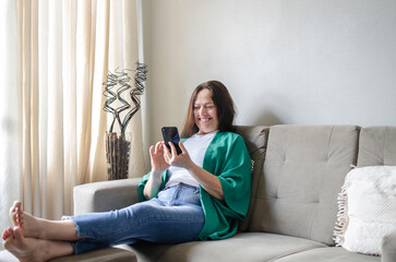 mulher de 50 anos sentada no sofá falando por chamada de vídeo no celular emocionada e sorrindo
