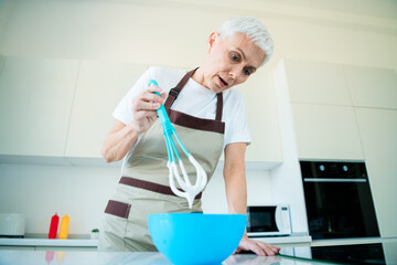 Senior woman in kitchen whisking ingredients in bowl, preparing a delicious homemade dish with...