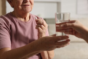 Caregiver giving glass of water to senior woman with medical pill indoors, closeup