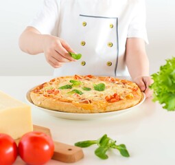 The boy puts on a pizza basil leaves; cheese, two tomatoes, lettuce are in front