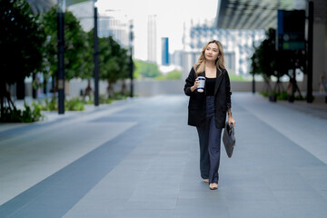 Stylish businesswoman walking in a modern urban setting with a coffee cup, exuding confidence, professionalism, and a dynamic career lifestyle in a sleek architectural city.