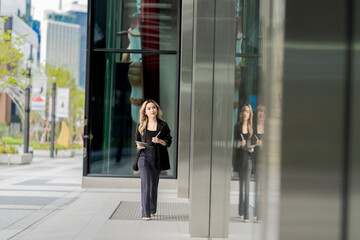 Confident businesswoman walking through a modern urban environment with reflective glass buildings, holding a tablet. A dynamic and empowering portrayal of contemporary professional life.