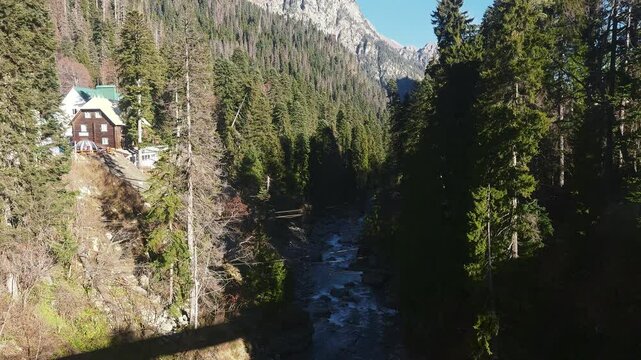 Stormy river flowing from mountains is located in canyon between tall trees and wooden buildings on hillside. Picturesque nature with cottages situated in remote area, view from above