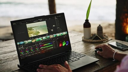 Video editor works on color grading footage at beach cafe. Man adjusts video clips on laptop with ocean view at sunset. Creative professional edits film at seaside, freelance lifestyle. Slow motion. - Powered by Adobe