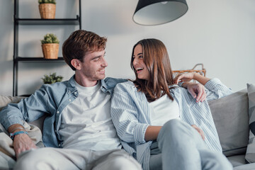 Young happy couple having fun talking laughing relaxing at home on couch, boyfriend embracing...