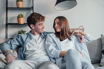 Young happy couple having fun talking laughing relaxing at home on couch, boyfriend embracing...