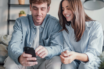 Happy millennial couple sit relax on couch in living room watching video on cellphone together, smiling young husband and wife rest on sofa at home browsing Internet using modern phone device.