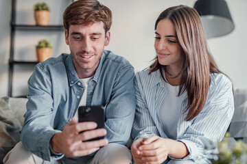 Happy millennial couple sit relax on couch in living room watching video on cellphone together, smiling young husband and wife rest on sofa at home browsing Internet using modern phone device.
