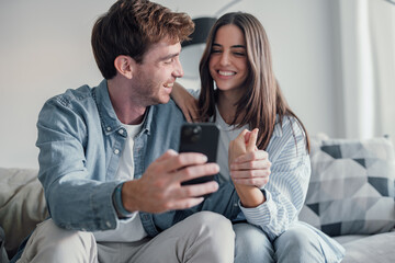 Fototapeta premium Cute couple greeting having fun video calling long distance friends and family catching up..Young man holding phone with girlfriend sitting on the sofa laughing and enjoying technology