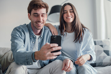 Cute couple greeting having fun video calling long distance friends and family catching up..Young man holding phone with girlfriend sitting on the sofa laughing and enjoying technology