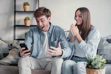 Excited millennial husband and wife relax on couch in kitchen feel euphoric win lottery online on...