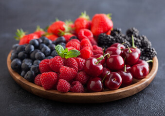 Fresh organic summer berries mix in round wooden tray on black kitchen table background. Raspberries, strawberries, blueberries, blackberries and cherries.