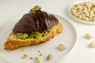 Croissant stuffed with pistachio paste and kadaif dough covered with dark chocolate on a white plate. Unusual pastry with delicate filling. Trend food concept. Selective focus.
