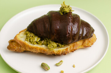 Croissant stuffed with pistachio paste and kadaif dough covered with dark chocolate on a gray plate on a green background. Unusual pastry with a delicate filling. Trend food concept. Selective focus.