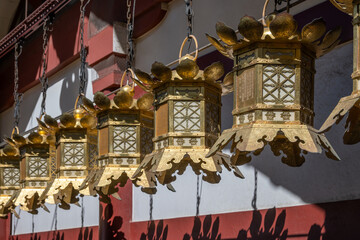 Ornate gold lanterns hang from chains, casting intricate shadows on a red and white temple wall in bright sunlight at Shitenno-ji Temple Osaka, Japan