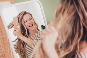 Young woman with blond hair smiling while brushing hair in living room mirror
