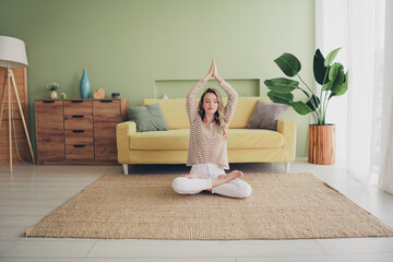 Young woman enjoying a peaceful weekend at home in casual style, meditating in a cozy living room...