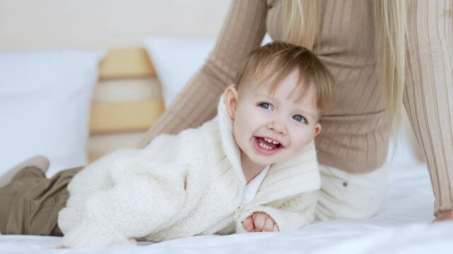 Cute smiling Caucasian baby boy wearing warm sweater lies on his belly. Mom sitting behind tickles her son.