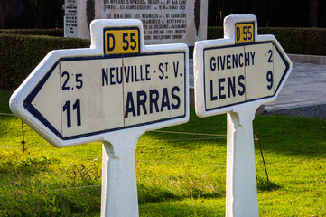 Road signs, from WI, distances and directions to Neuville-St. V. and Arras, and Givenchy and Lens. A memorial plaque in the background about fallen soldiers, the location is a war memorial or cemetery