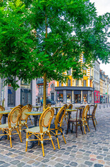 Street restaurant with wooden tables and chairs on cobblestone street and green tree, old colorful buildings in Lille city center, vertical view, French Flanders, Nord department, Northern France