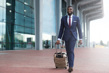 Portrait of young african american man walking in airport terminal with luggage, smiling african...