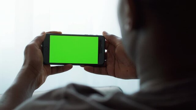 An African American man holds a smartphone with a bright green screen while sitting at home. He appears absorbed in the device, highlighting a modern digital lifestyle.