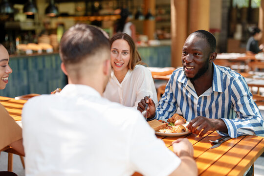 A diverse group of friends, including men and women, share a leisurely breakfast at an outdoor cafe. They are smiling and wearing casual attire, enjoying each other's company on a sunny day.