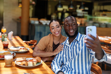 Young adult Black male and multiracial female couple capture a joyful selfie while enjoying breakfast with diverse friends at a sunny café. The warm atmosphere highlights their happiness.