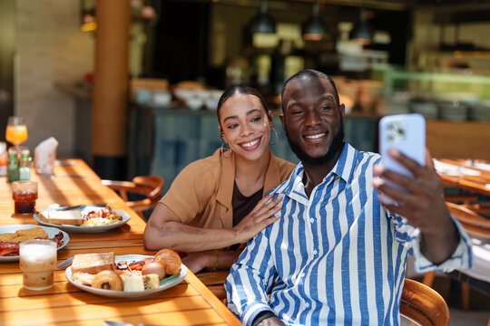 Young adult Black male and multiracial female couple capture a joyful selfie while enjoying breakfast with diverse friends at a sunny café. The warm atmosphere highlights their happiness.