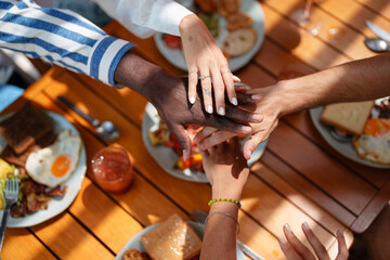 A diverse group of friends gathers around a sunlit table, stacking hands above a nutritious breakfast. Plates feature eggs, toast, and fresh juice, highlighting camaraderie and healthy eating.