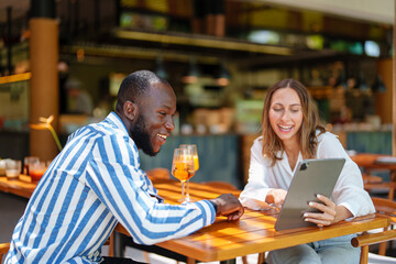 A young Black male and Caucasian female entrepreneur engage in a lively discussion over a digital tablet at a vibrant tropical bar. They appear enthusiastic and focused.