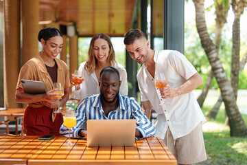 A diverse group of young adult entrepreneurs engages in collaborative work on a laptop and digital tablet at a tropical bar. The scene conveys teamwork, innovation, and creativity.