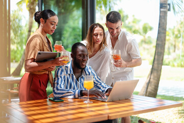 A group of diverse young adult entrepreneurs, including both men and women, work together in a tropical bar. They are engaged with a laptop and digital tablet, enjoying drinks.