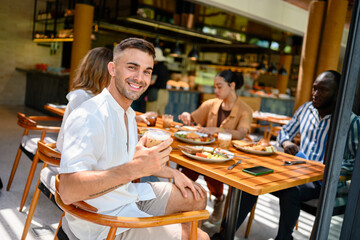 A young Caucasian male enjoys breakfast with friends of diverse ethnicities in a cozy cafe. The relaxed atmosphere features warm lighting, casual attire, and delicious food.