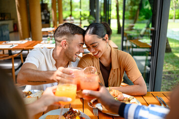 A Caucasian male and multiracial female couple enjoy brunch with diverse friends in a cozy bar, laughing and toasting with fresh juice. The cafe’s bright, sunlit interior offers a warm atmosphere.