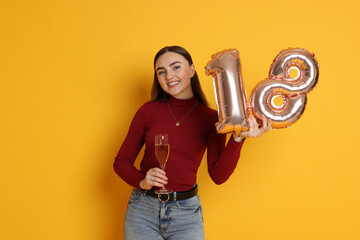 Coming of age party - 18th birthday. Happy young woman with number shaped balloons and glass of sparkling wine on yellow background