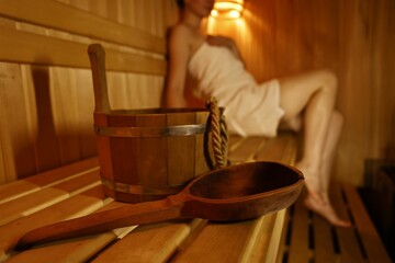 Bucket with ladle and woman on wooden bench in sauna, selective focus