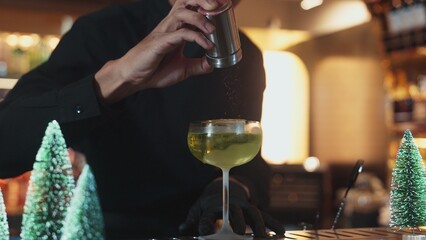 Cropped bartender garnishing caramelized citrus slice on cube ice in green Midori Sour cocktail served in stylish stemmed glass. Perfect for sophisticated bar settings christmas tree theme. Vinosity.