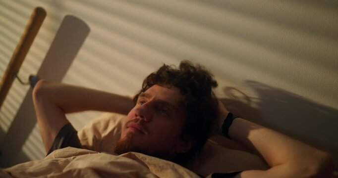 A brunette man with curly hair sets his alarm for a late time and continues to rest in his bed in the morning