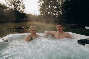 A young couple of a man and a woman are happily relaxing in a jacuzzi on a weekend in the mountains. The background is a beautiful landscape of nature.