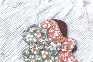 Reusable cloth menstrual pads in green, pink, and brown with cherry blossom floral print, made of muslin fabric, placed on a textured fabric background concept of eco-friendly alternatives