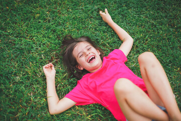 Cheerful young girl 7 years old in a bright pink t-shirt lying on green grass and laughing...