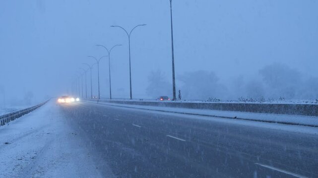 Cars riding on the snowy highway with headlights turned on, amid heavy winter weather conditions. Car traffic during snowstorm conditions on a highway.