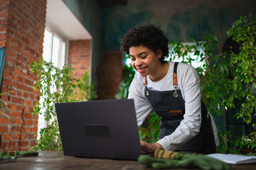 African woman florist gardener working in botanical store using laptop order plants communicating...