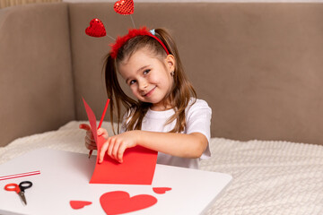 Cute little girl in head rim with red hearts makes Valentine's Day cards using colored paper,...