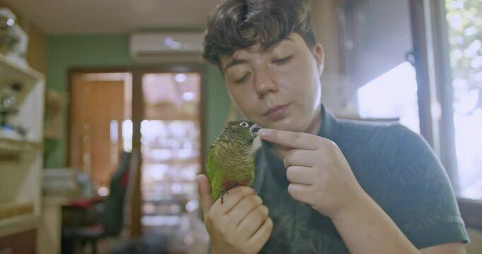 A young person gently pets and cuddles their pet green-cheeked conure parrot at home. They share a loving bond, showing affection and care.