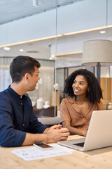Two young diverse coworkers business people using laptop and talking at office meeting. Busy happy team professional employees working together on computer discussing online technology data at work.