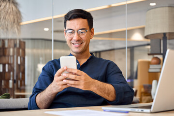 Smiling busy professional Hispanic business man sitting in office holding mobile cellphone. Young...