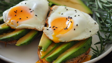 Close Up Two Toasts avocado fried egg Spreading Yolk, rosemary. Preparing healthy toast breakfast avocado slices rye wholegrain bread, sunny side up egg on top. Healthy breakfast for two, couple
