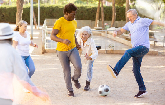 Joyful senior multinational friends playing soccer on a sandy playground on a warm spring evening